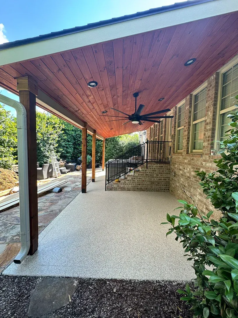 Covered stone patio with wood ceiling, ceiling fan, and steps beside a brick wall and garden greenery