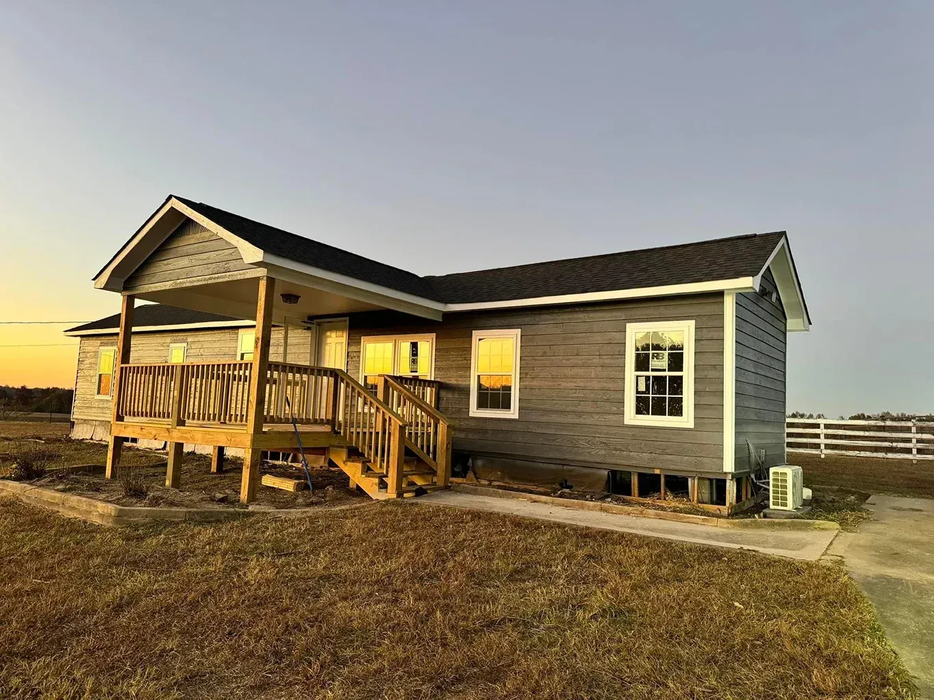 Single-story gray house with a wooden porch at sunset in a rural field