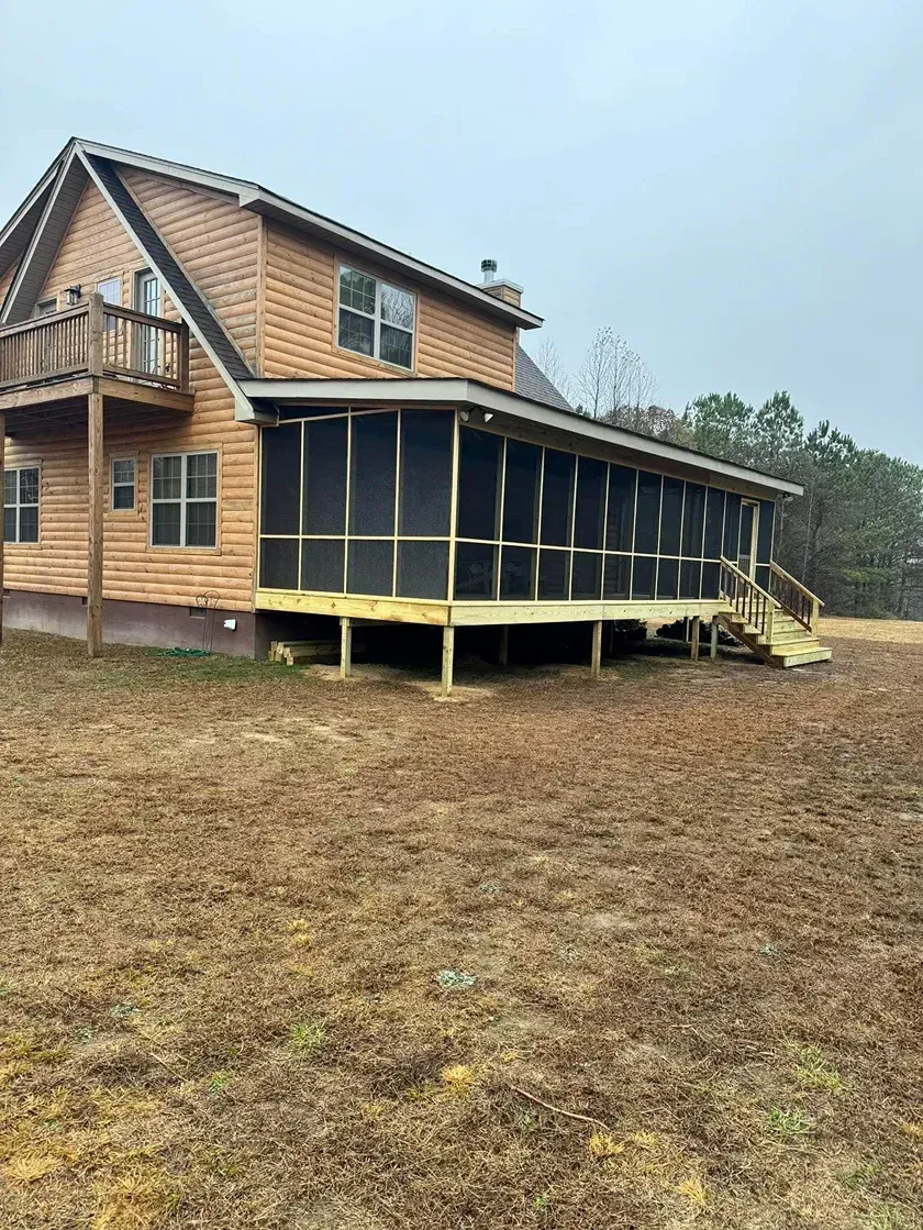 Wooden house with a screened porch and balcony in a muddy yard under an overcast sky