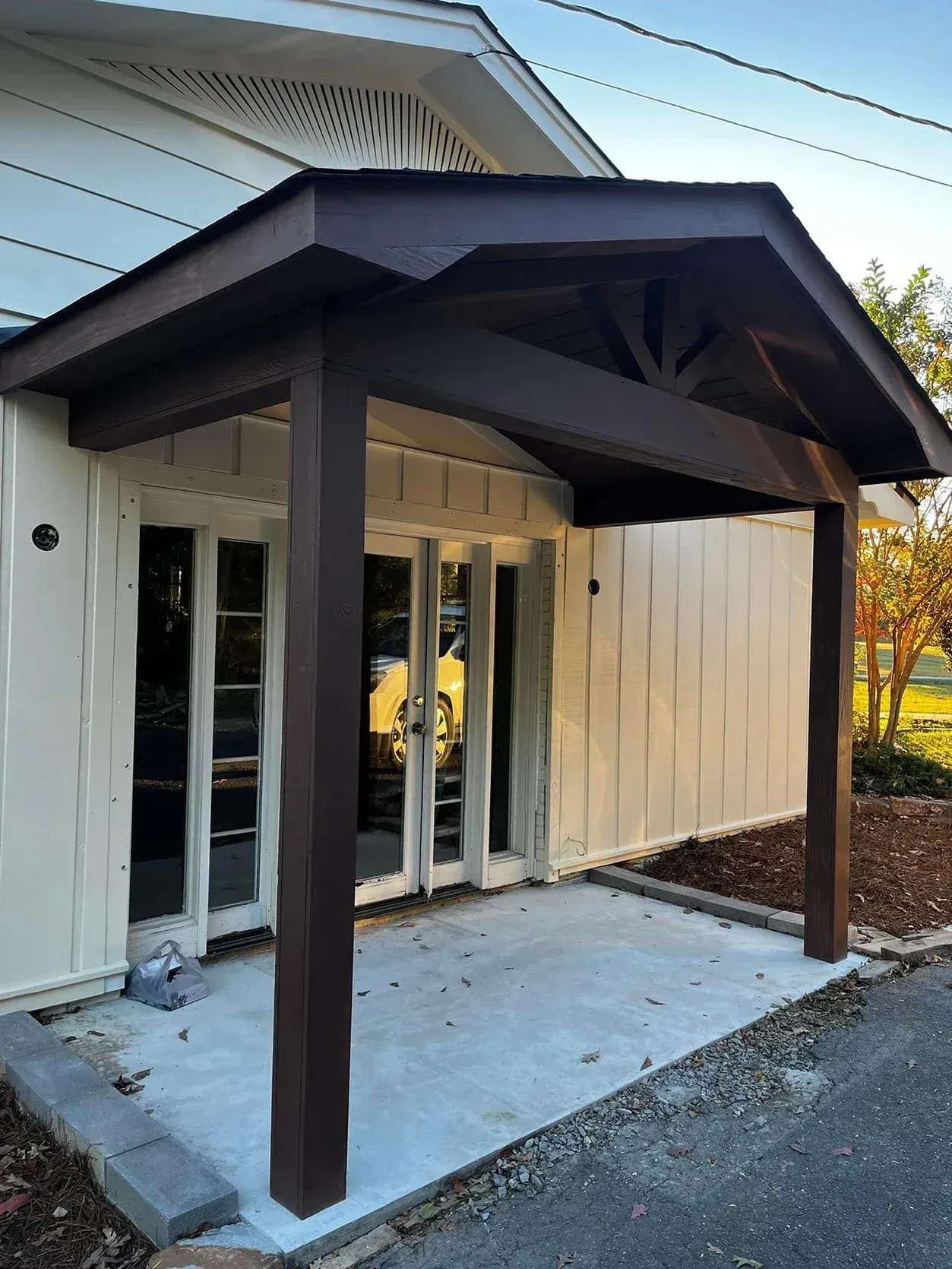 Covered front entry with dark wood posts and awning, white siding, and glass doors leading into a home