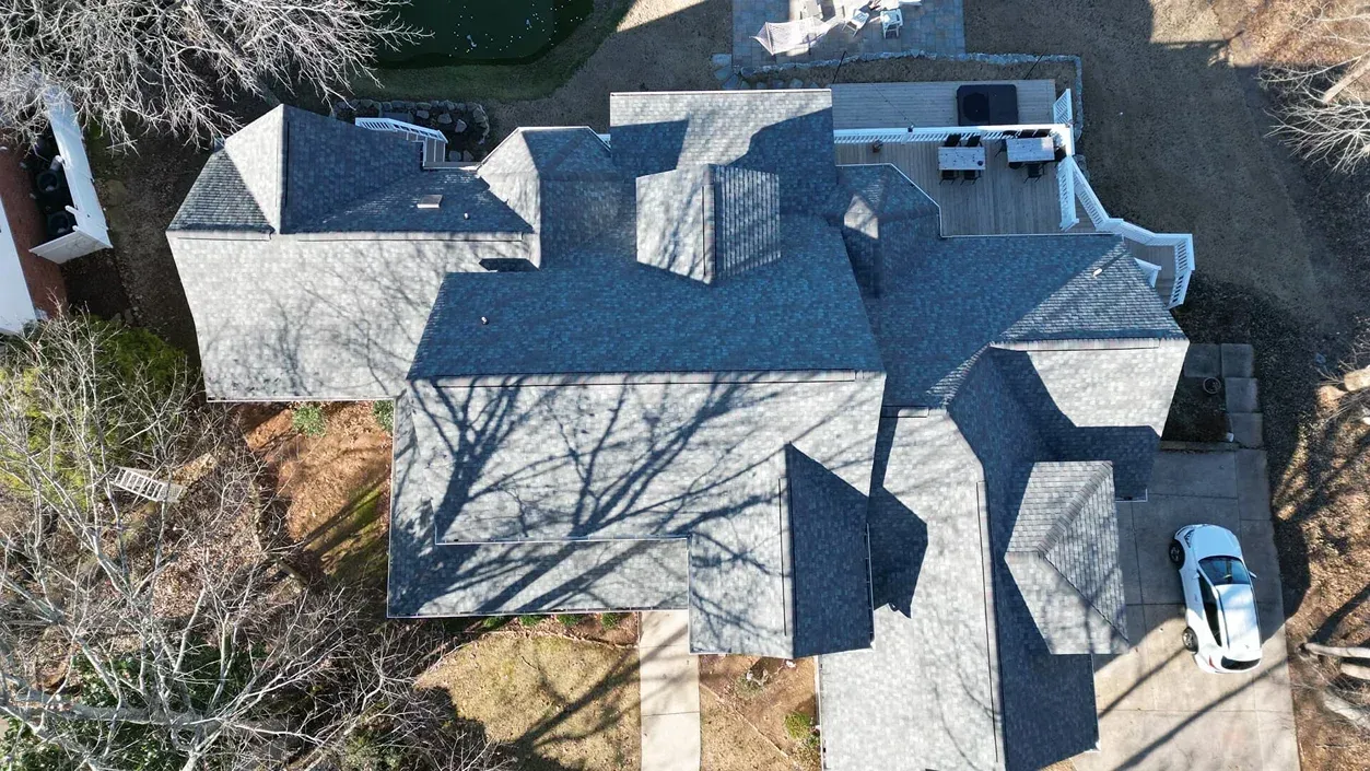 Aerial view of a gray-shingled house roof with dormers, chimneys, and a driveway with two parked cars.