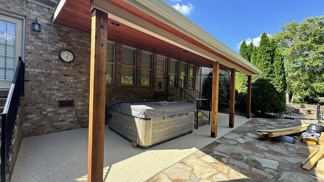 Covered patio beside stone house with hot tub, wooden posts, and trees in a sunny backyard