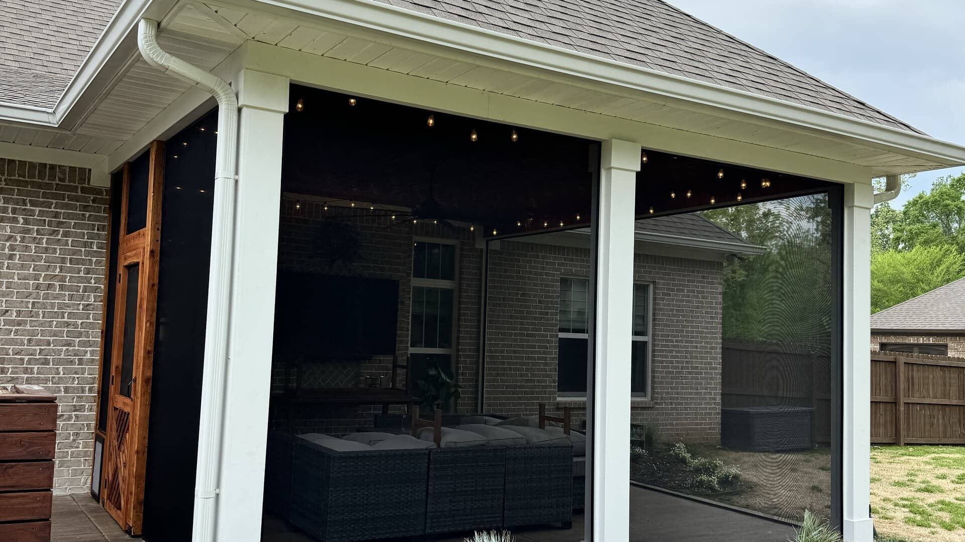 Covered backyard patio with white columns, dark wicker furniture, and string lights under a roof overhang.
