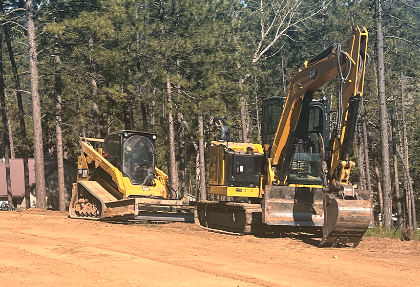 Yellow excavator and skid steer parked on a dirt lot with tall trees in the background.