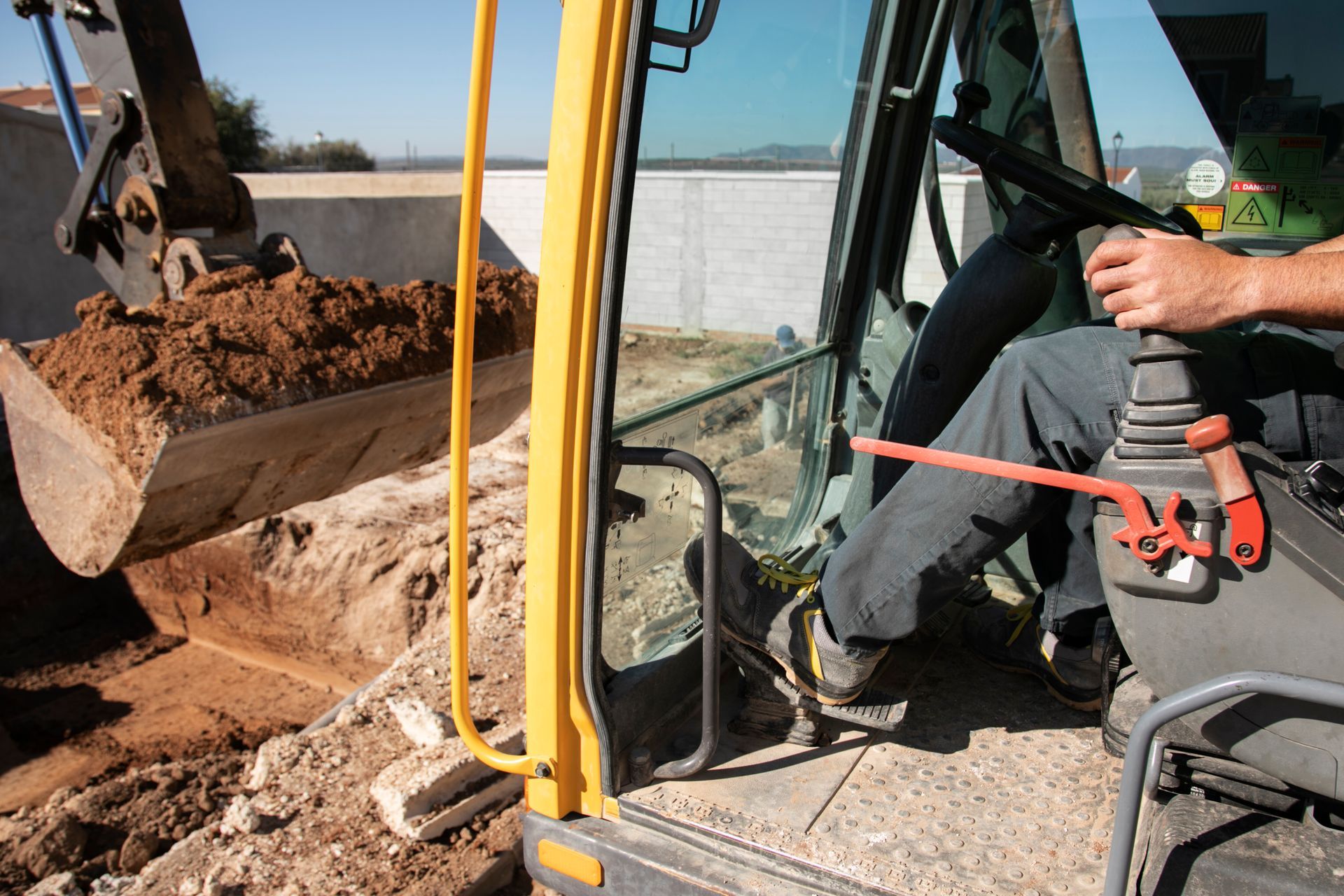 Excavator operator using controls to move a scoop of dirt. The machine is yellow and black, in an outdoor setting.