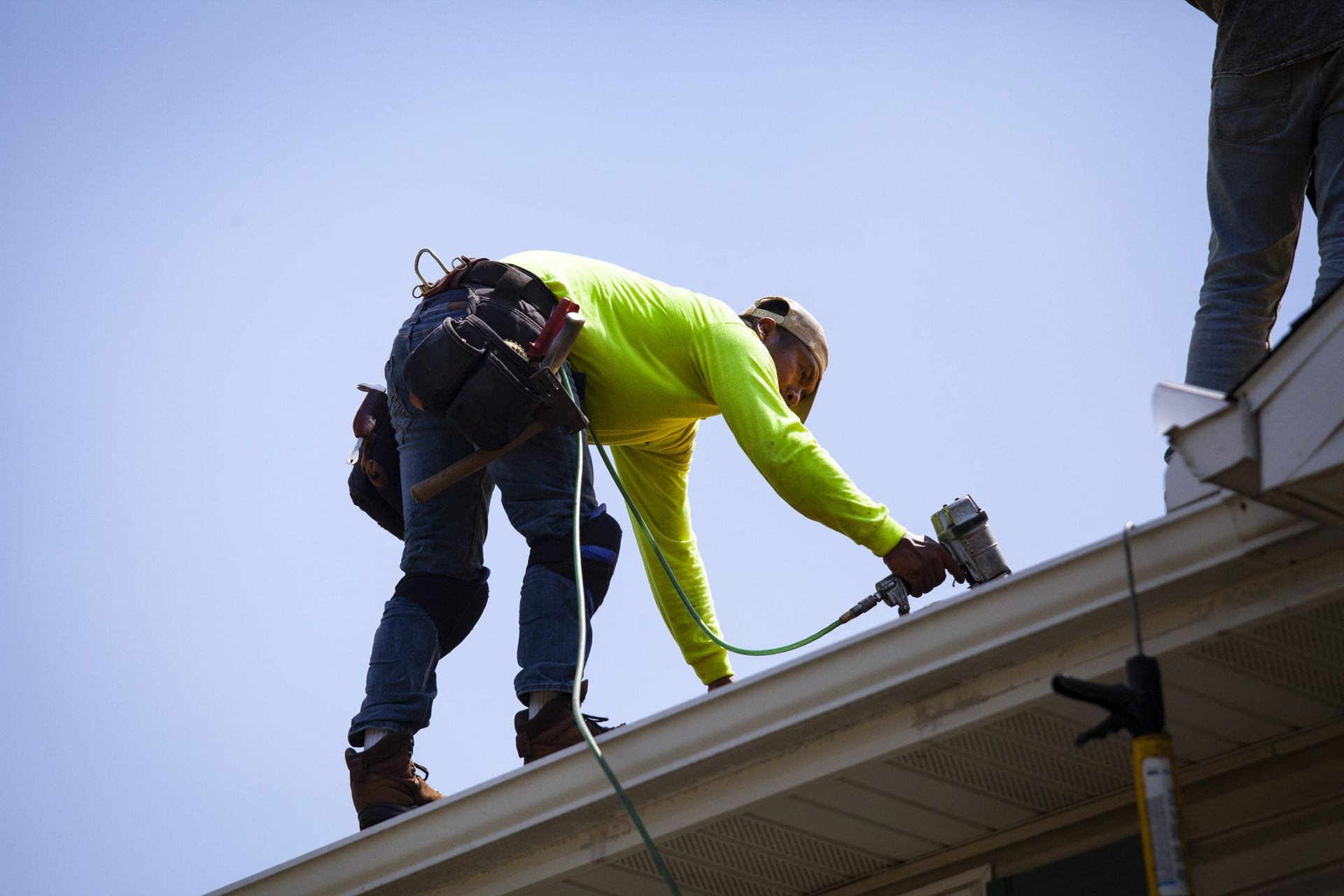 A roofer in a bright yellow long-sleeved shirt uses a nail gun while working on a roof against a clear blue sky.