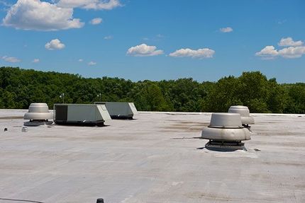 A flat roof with several HVAC units and ventilation fans against a backdrop of trees and a bright blue sky.