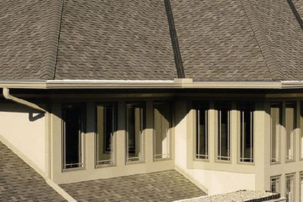 Exterior view of a house with grey shingled roofing, light tan walls, and a row of tall, multi-pane windows.