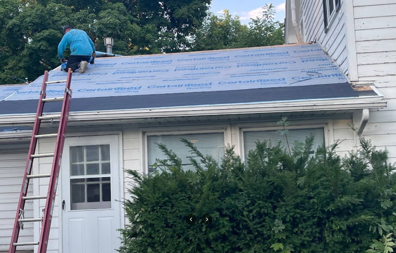 A worker in a blue shirt on a roof partially covered in blue underlayment, accessed by a red ladder at the side of a house.