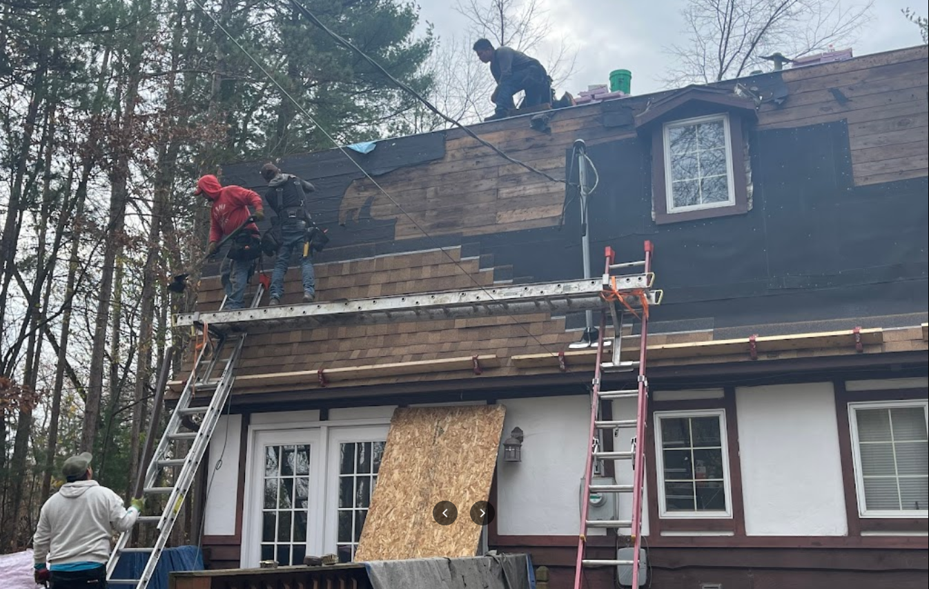 Workers install siding on the upper story of a house with two ladders and scaffolding in a wooded setting.
