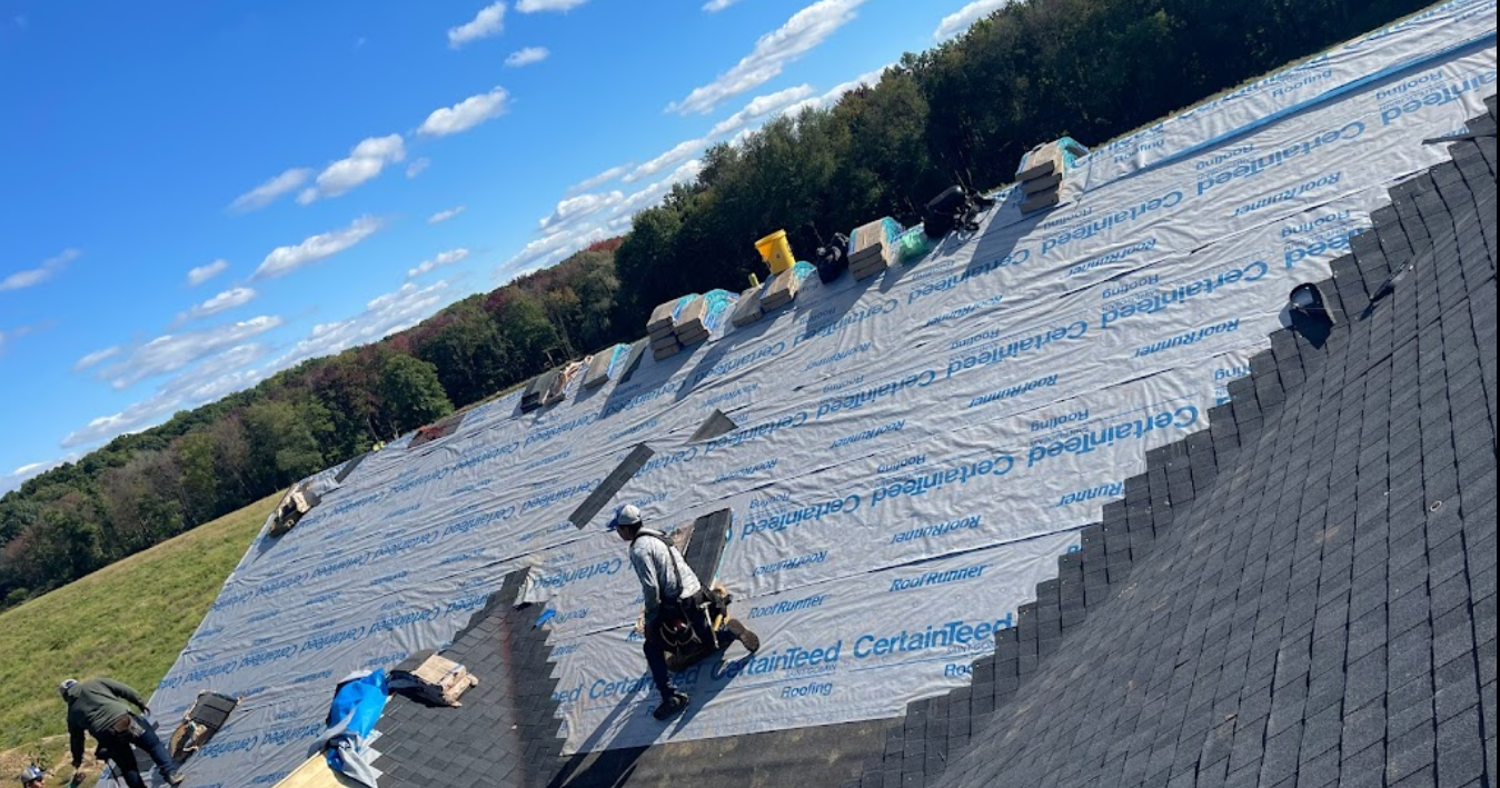 Workers install shingles on a roof covered in white underlayment against a blue sky with trees in the background.