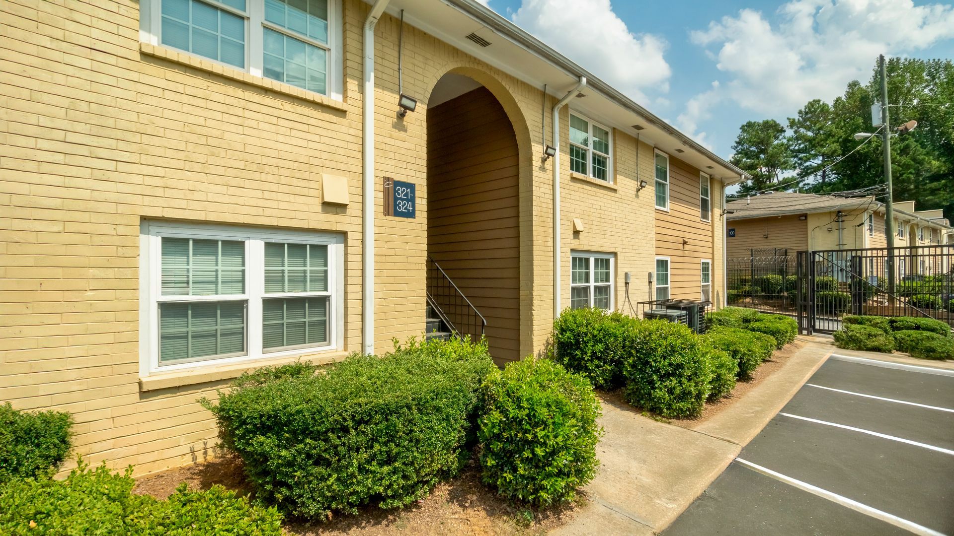 Brick apartment building with white-trimmed windows, arched entryway, shrubs, and parking area on a sunny day