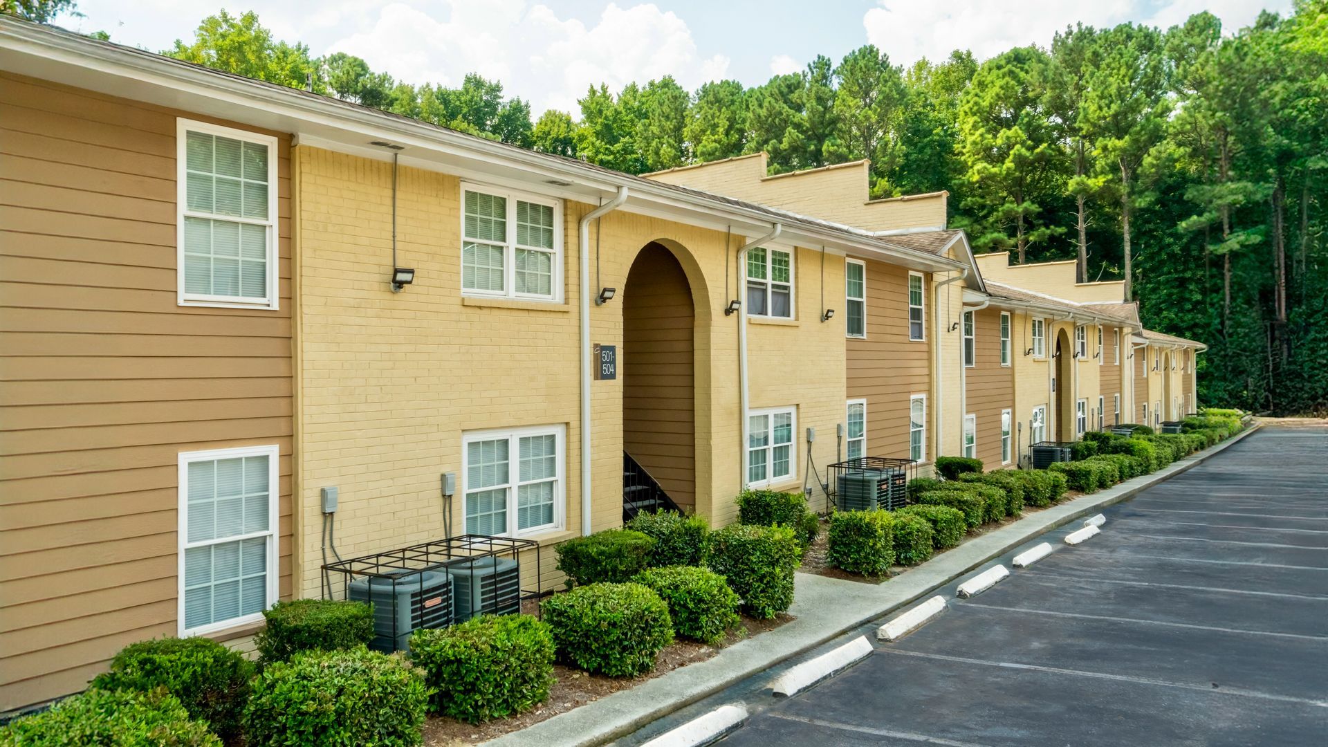 Row of beige apartment buildings with white-trim windows beside a paved parking lot and trees
