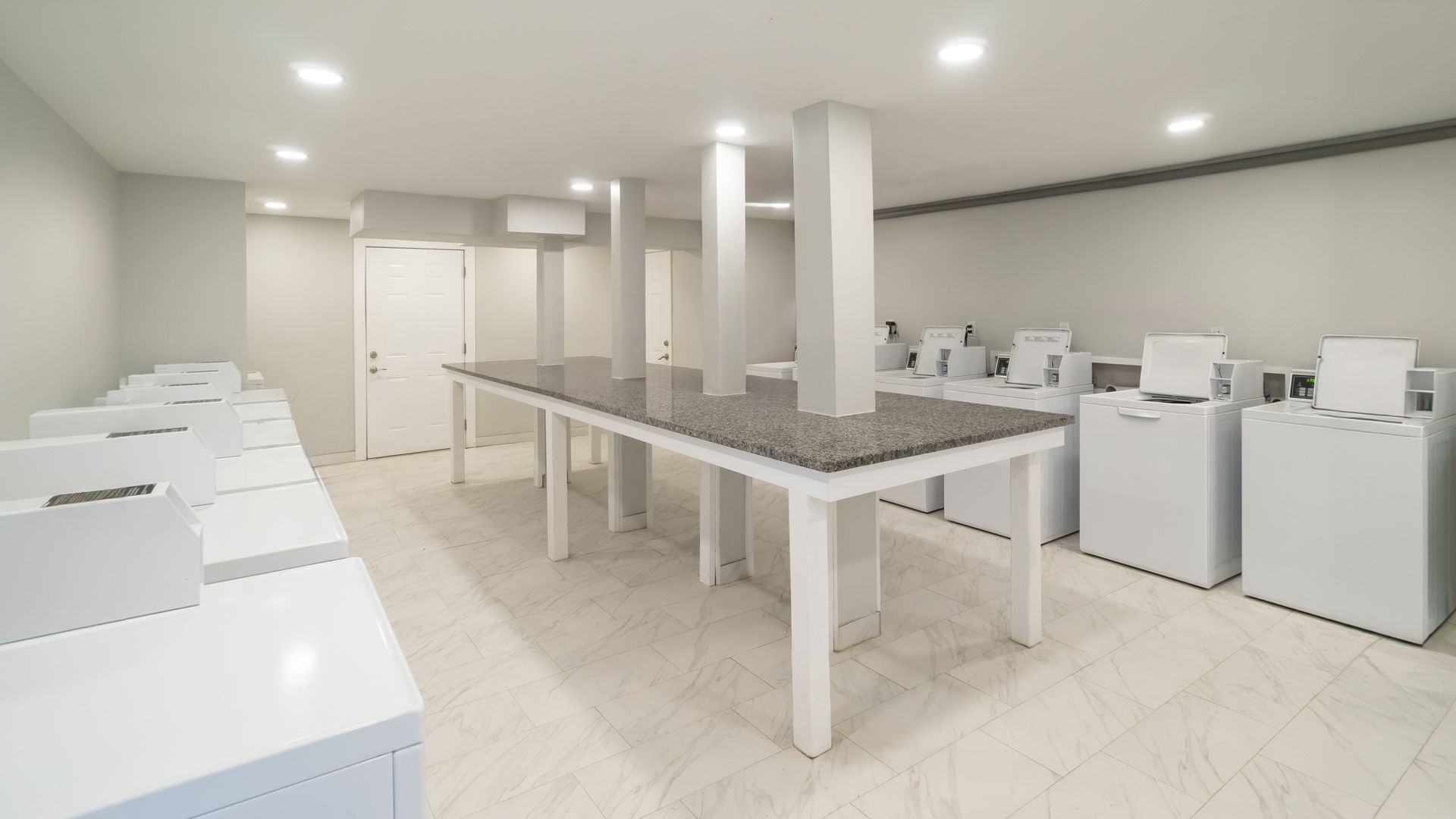 Bright, modern kitchen or laundry room with white appliances and long central tables under recessed lighting