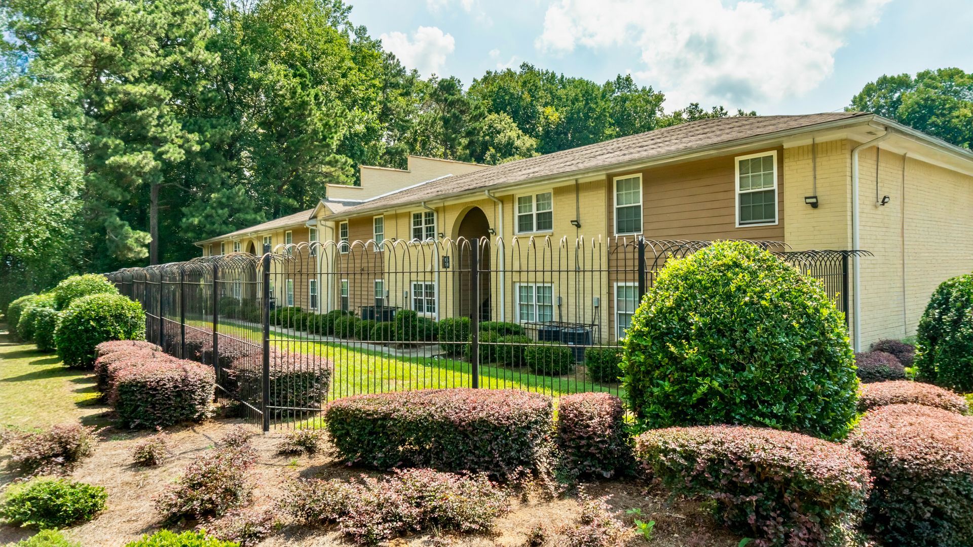 Single-story beige apartment building with black fence, manicured shrubs, and trees in front