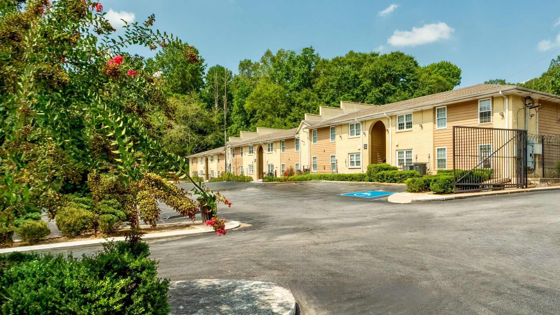 Stone apartment buildings along a paved road beside lush green trees on a sunny day