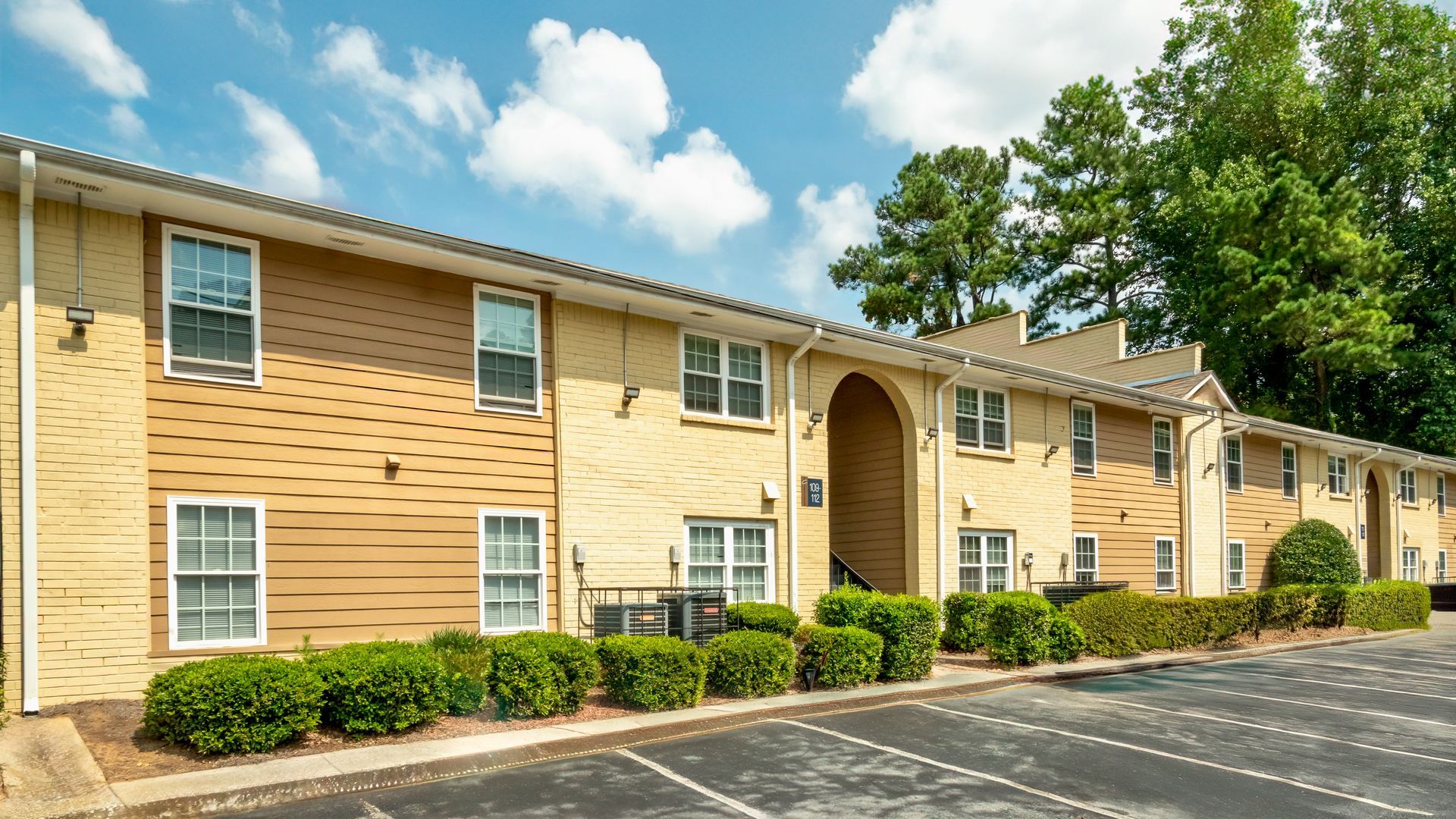 Apartment building exterior with tan siding, white windows, and trimmed shrubs under a partly cloudy sky