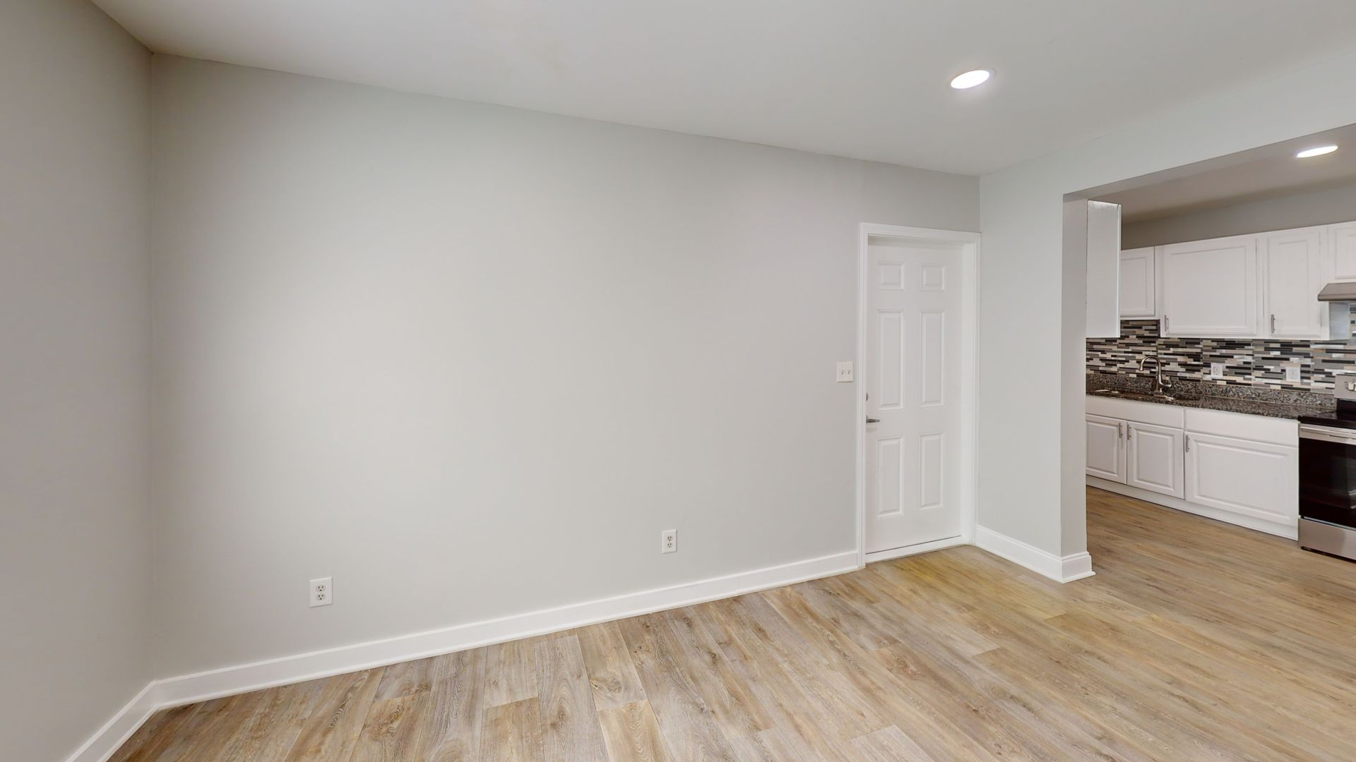 Empty living room with light wood floors, gray walls, and an open view to a kitchen area.
