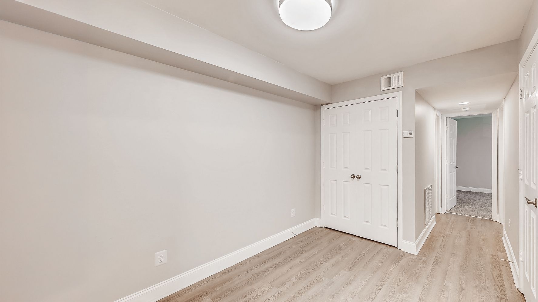 Empty white hallway with light wood floors, closet doors, and a doorway leading to another room