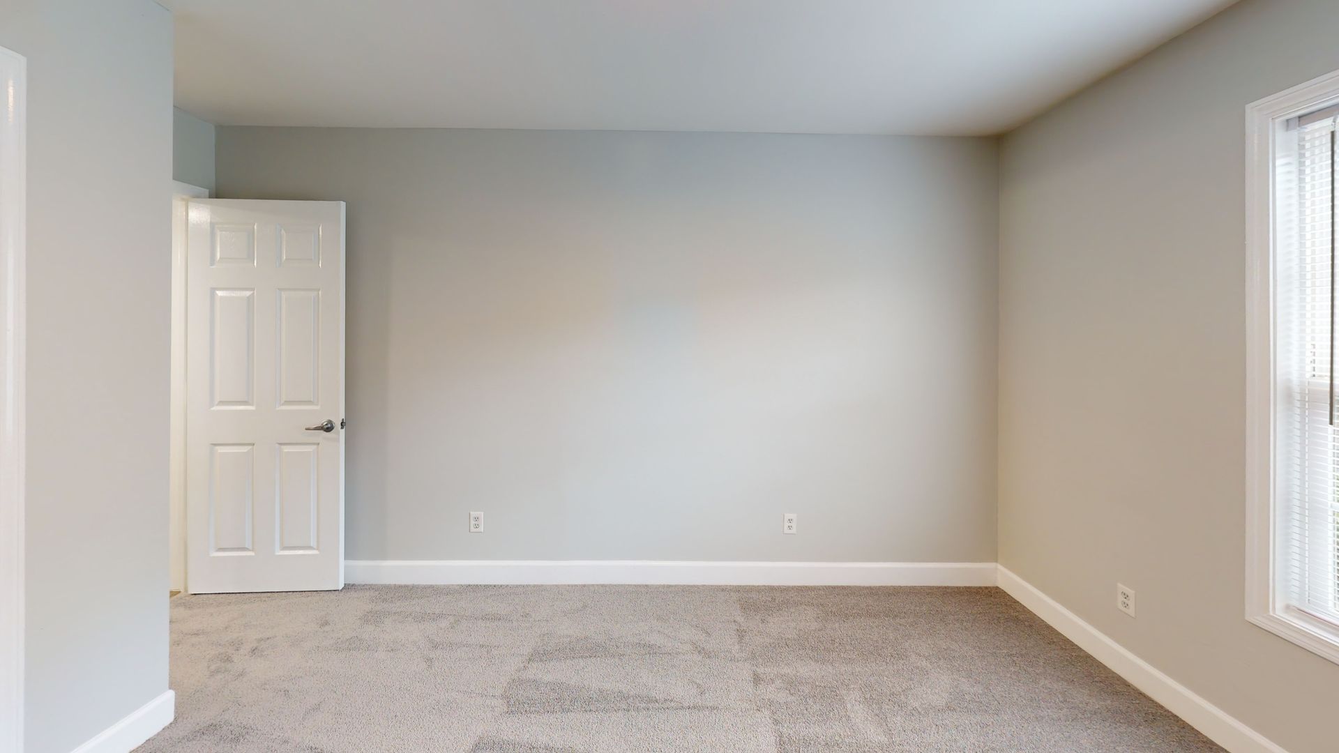 Empty beige bedroom with white door, gray carpet, and large window letting in light