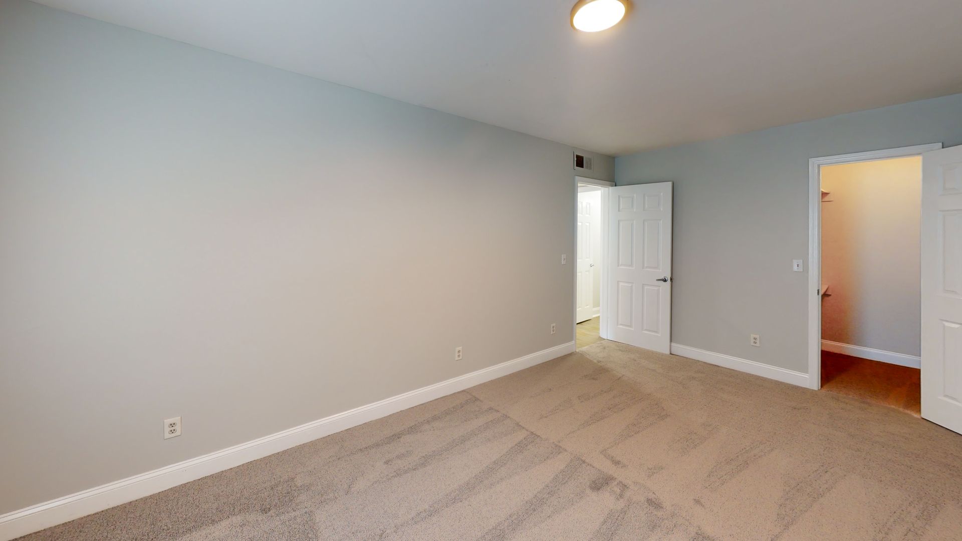 Empty beige bedroom with carpet, white closet doors, and an open doorway to a hallway.