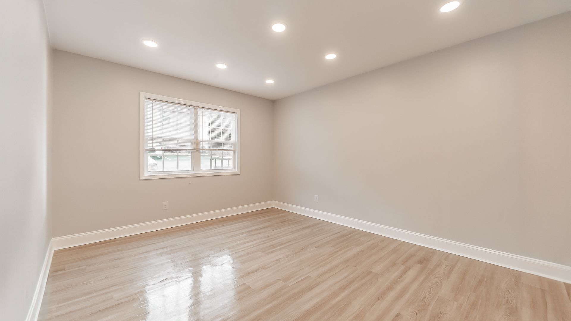 Empty beige living room with light wood floors, recessed lights, and a window.