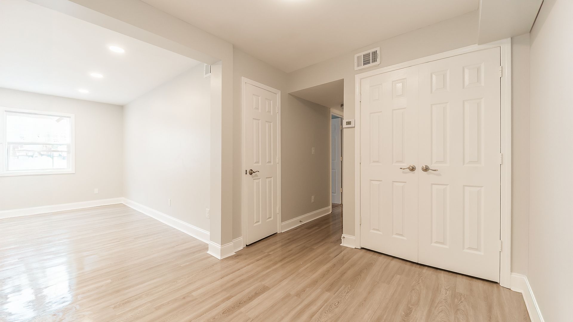 Bright empty hallway and living area with white walls, wood floors, and double closet doors