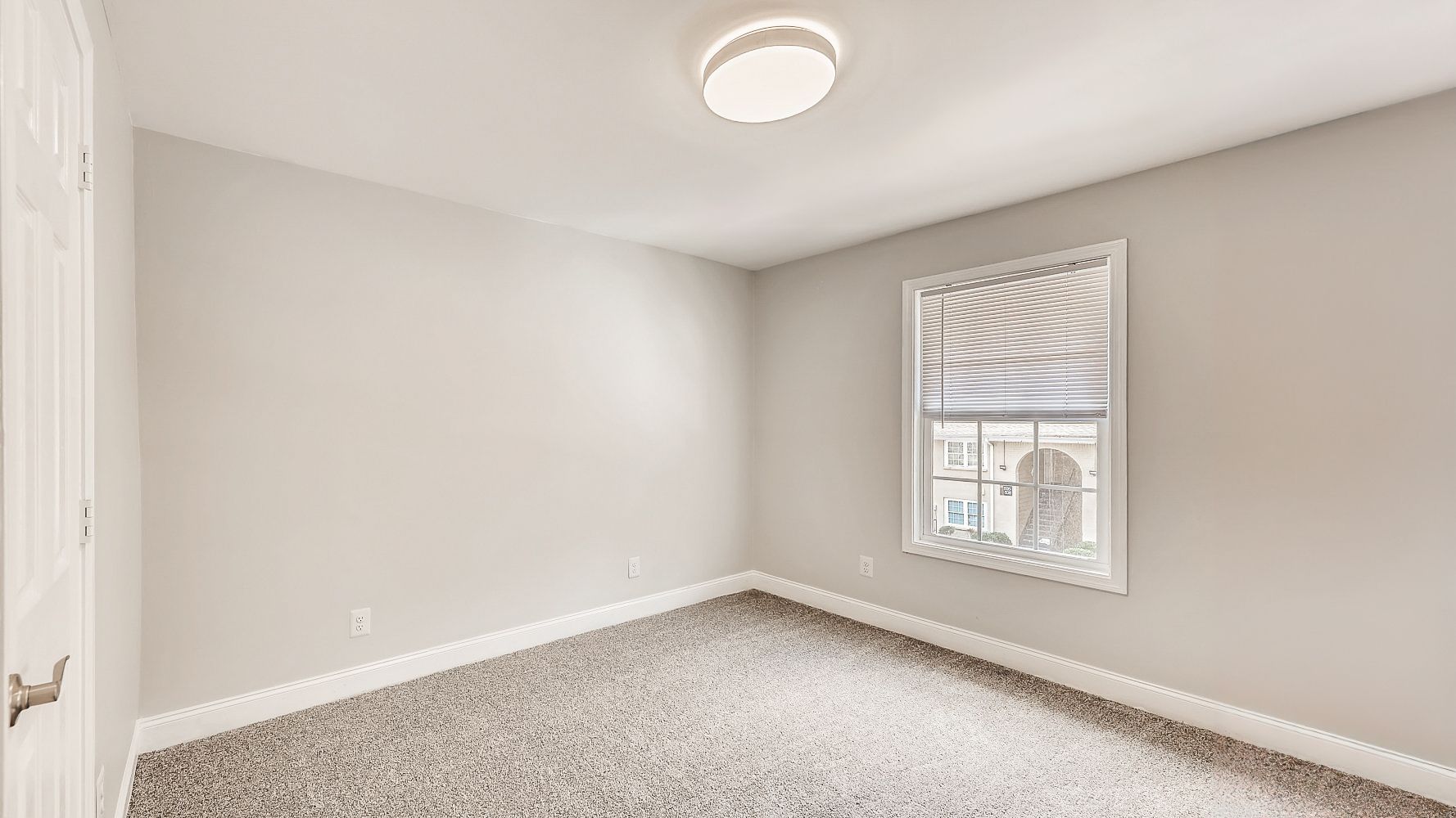 Empty light gray bedroom with carpet, a window with blinds, and a ceiling light fixture