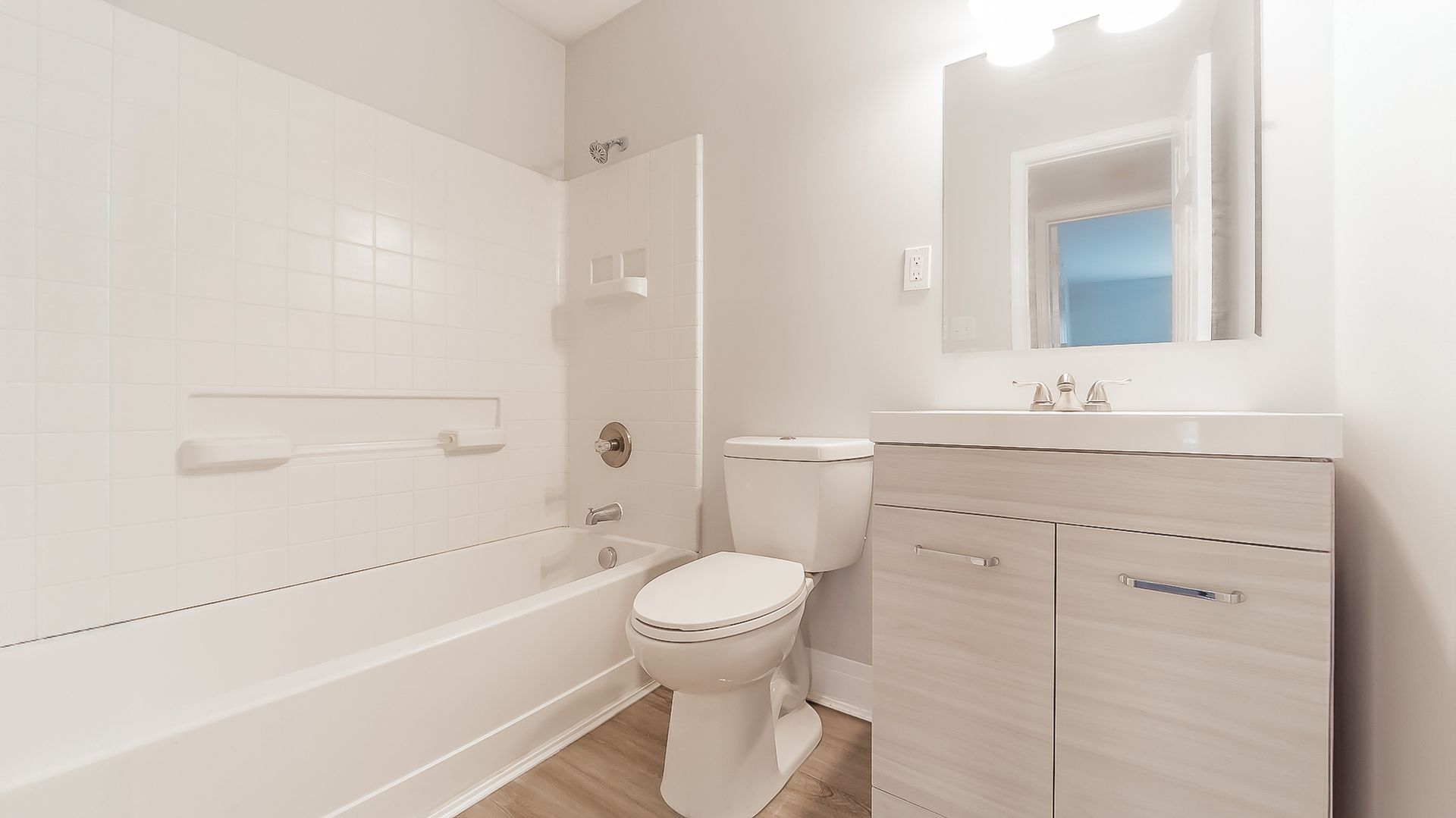Bright white bathroom with bathtub, toilet, vanity sink, mirror, and wood-look floor.