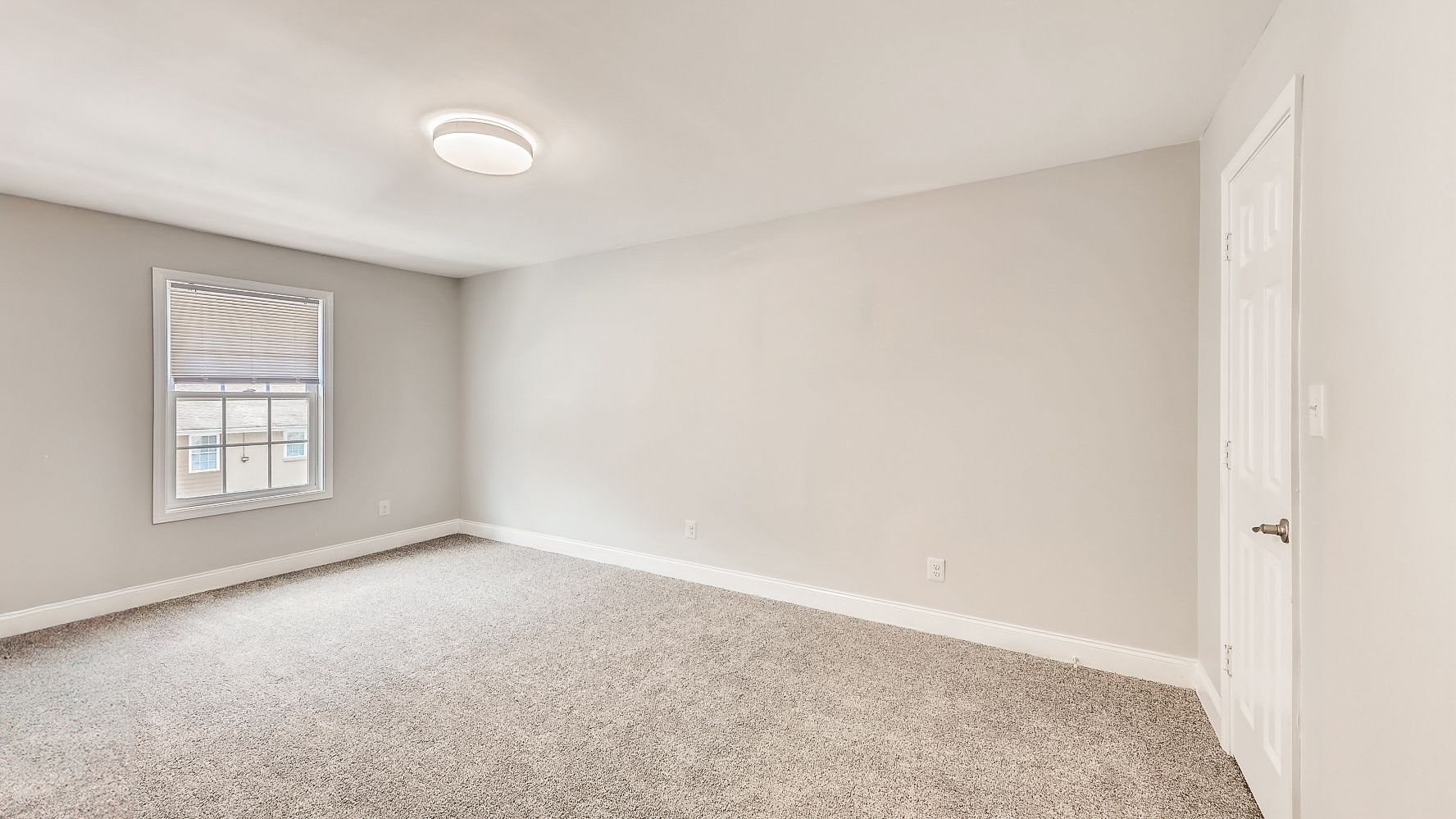 Empty beige bedroom with gray carpet, a window, and a white door.