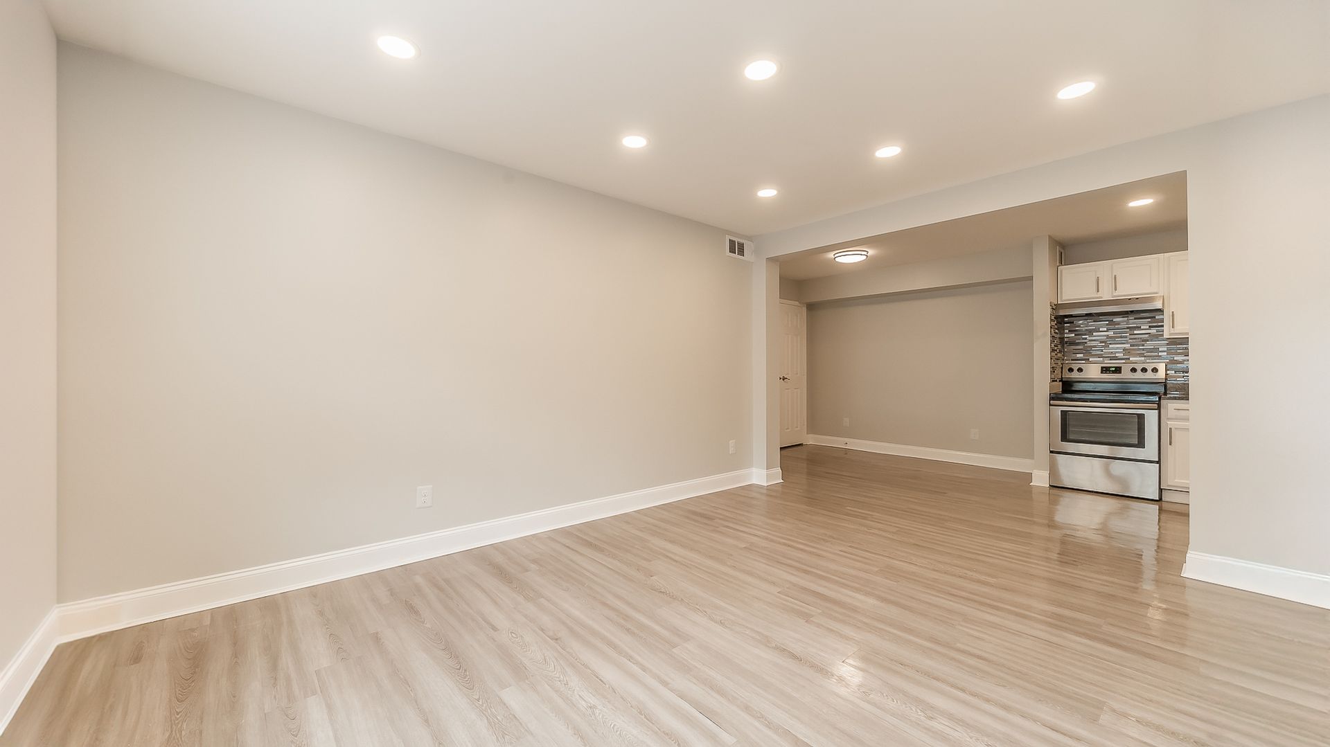 Empty modern living room with light wood floors, recessed lights, and open kitchen area in the background