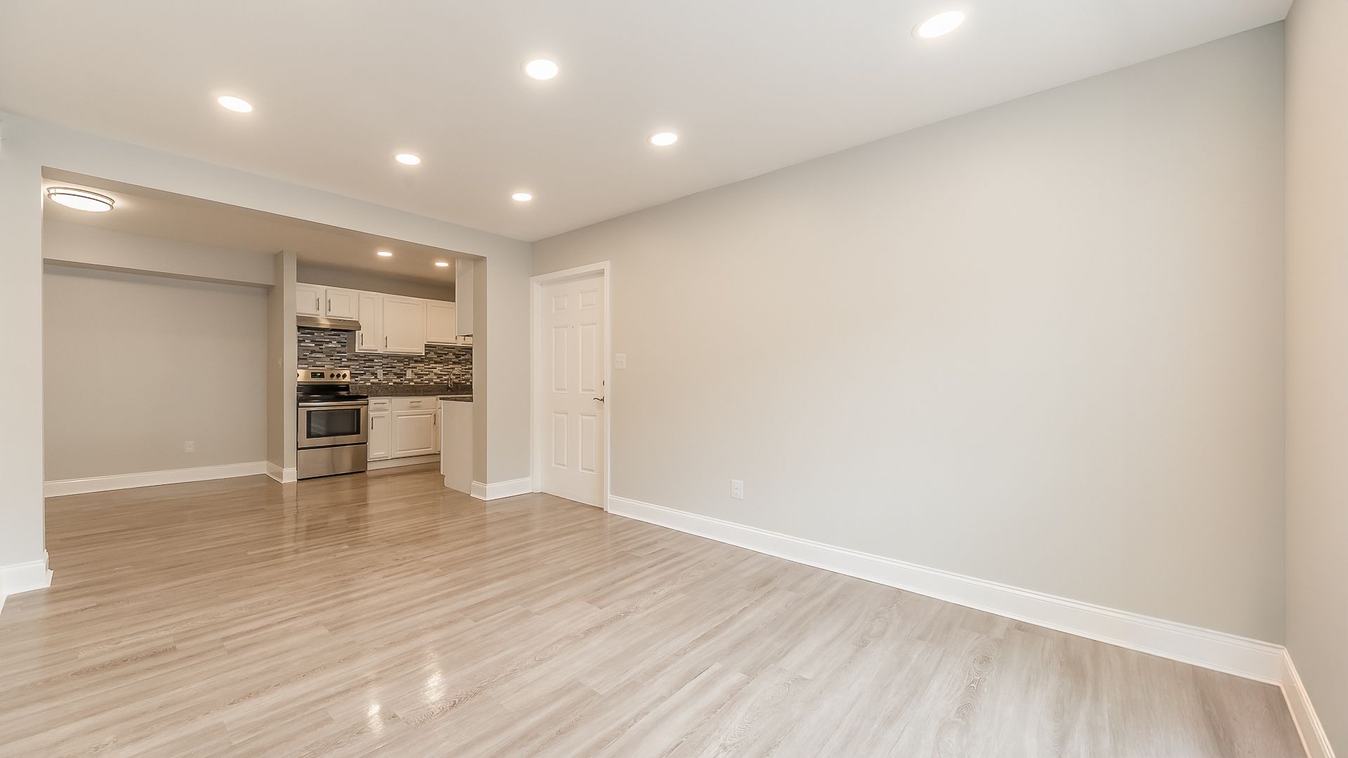 Empty modern living room with light wood floors, recessed lighting, and open view to a kitchen nook.