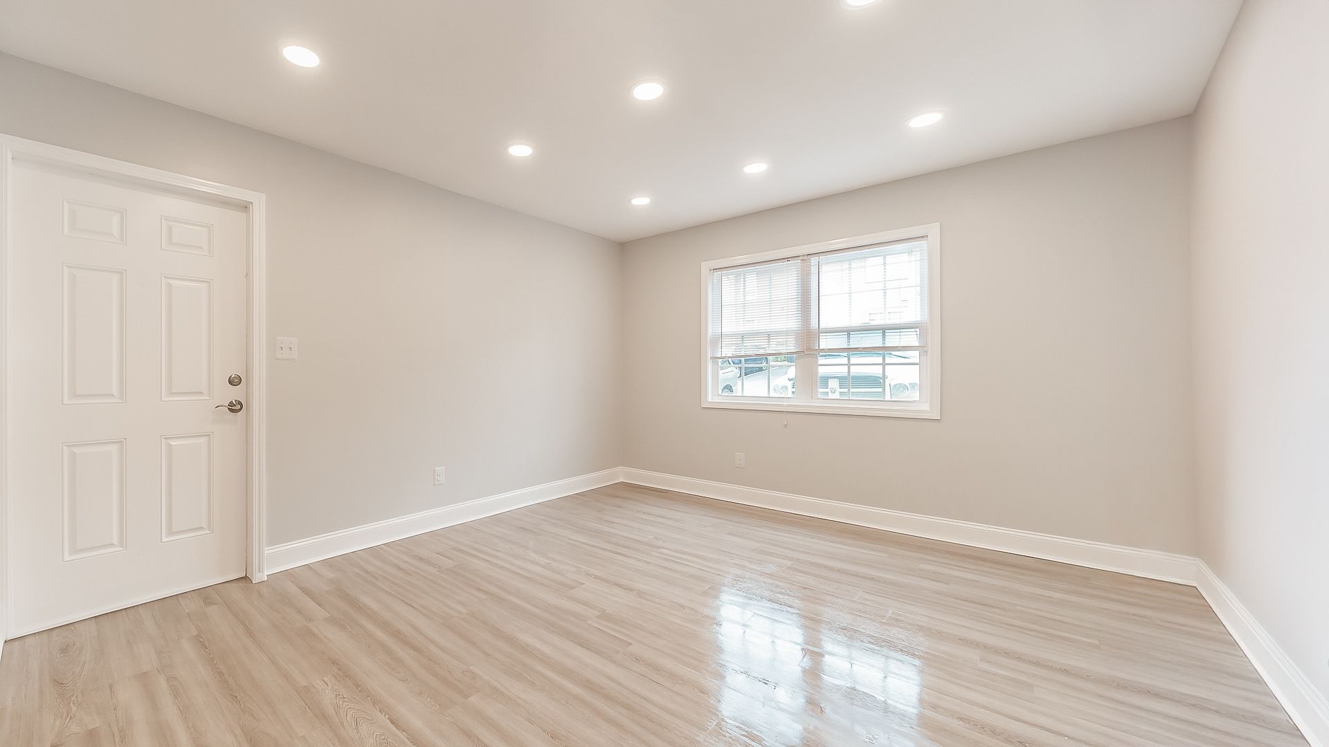 Empty beige room with wood-look floors, recessed lights, white door, and a small window.