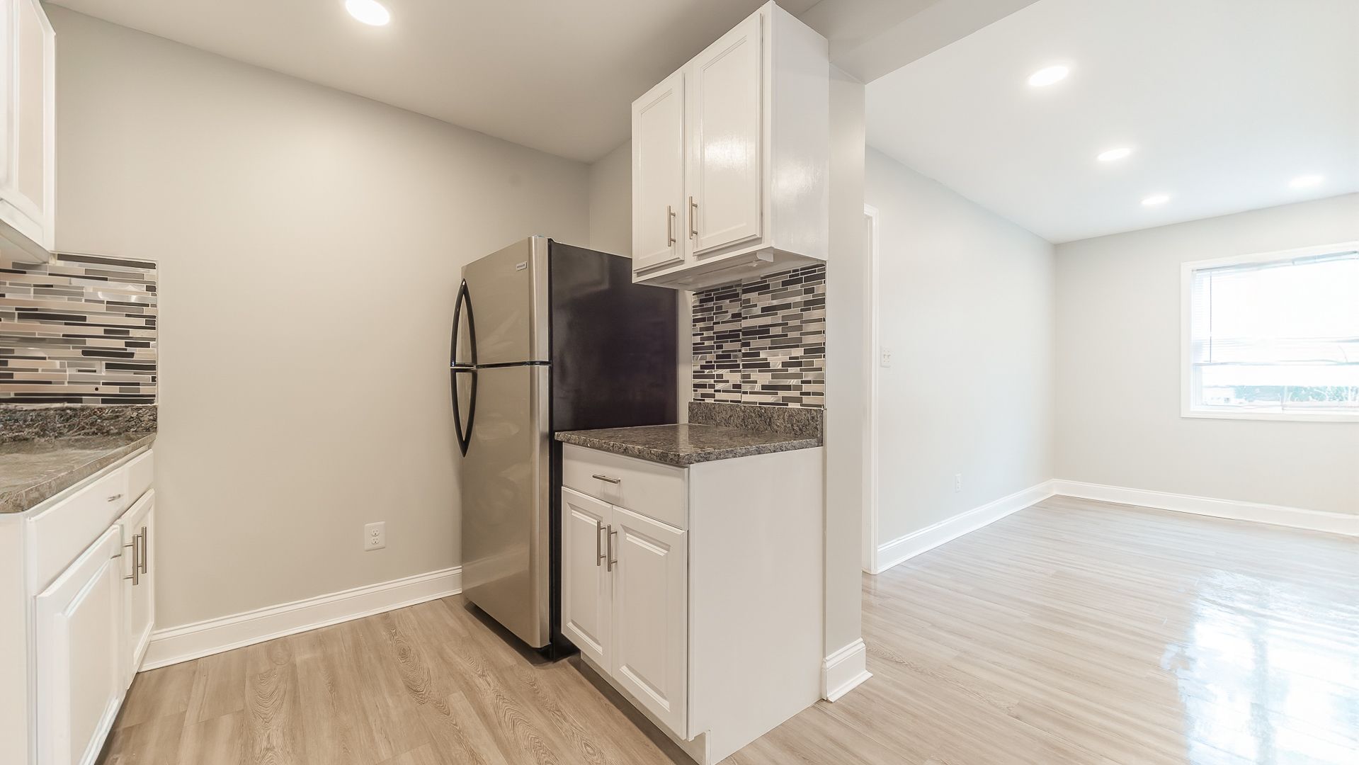 Bright modern kitchen with stainless steel fridge, white cabinets, and light wood floors opening to a sunlit room