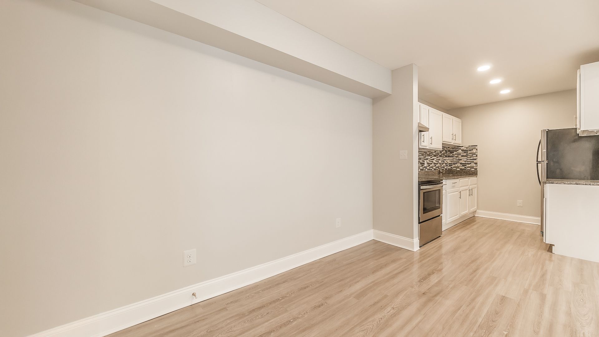Empty modern room with light wood floors, white walls, and a kitchen area in the back.