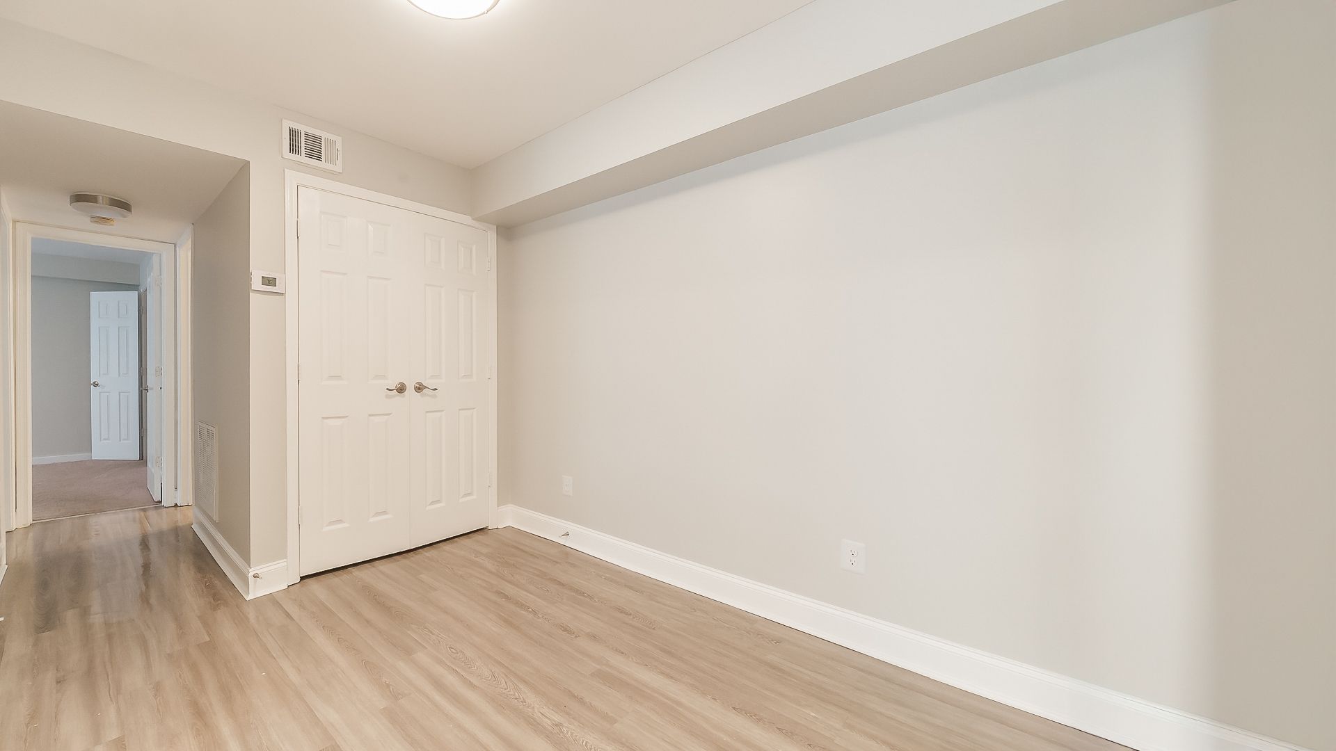 Empty white bedroom with light wood flooring, closet doors, and hallway visible through an open doorway