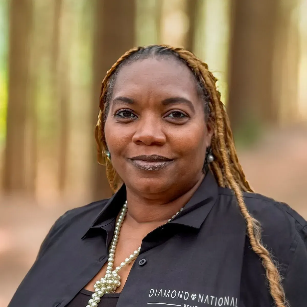 Portrait of a person outdoors in a black shirt with a pearl necklace, standing before a blurred wooded background