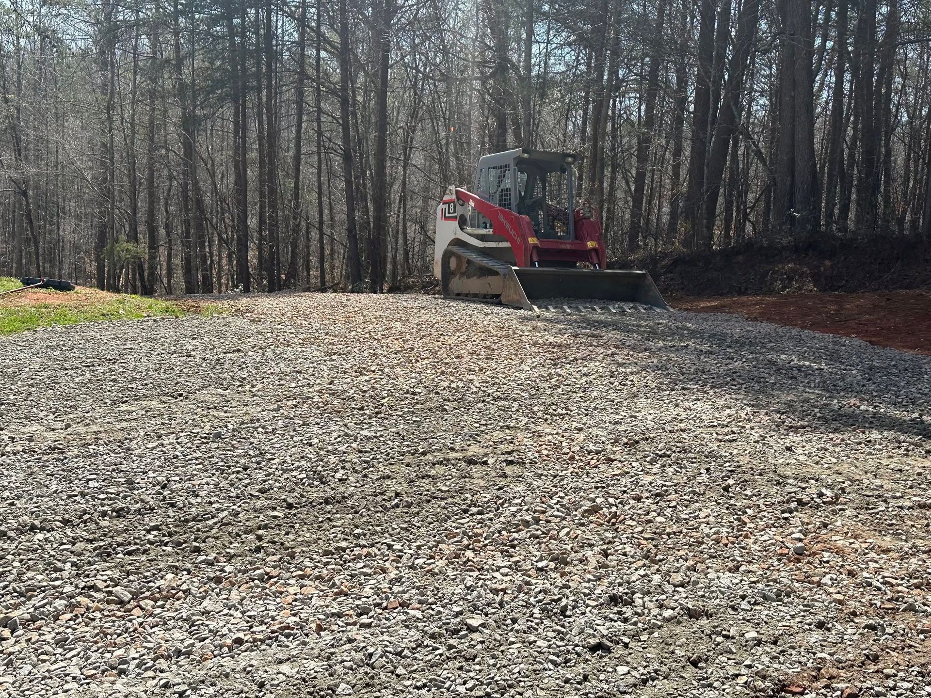 Red excavator on a gravel lot beside leafless woods in bright sunlight