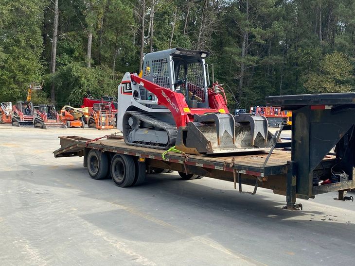 Red skid-steer loader on a flatbed trailer in a wooded lot with other equipment nearby