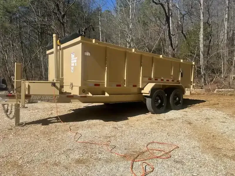 Tan dump trailer parked on a gravel lot beside woods, with hitch and dual wheels visible
