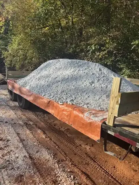 Large pile of gray gravel on an orange tarp beside a dirt road, with trees in the background