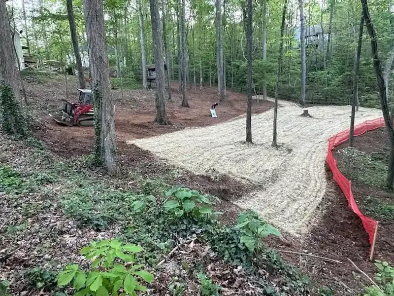 Wooded dirt trail under construction with a small red excavator and pale gravel path edged by red tape