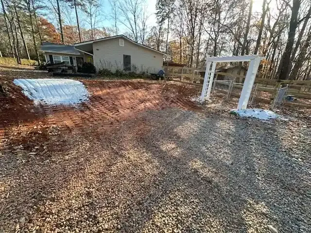 Gravel driveway beside a house with a white frame structure and a low, snow-covered mound, surrounded by bare trees