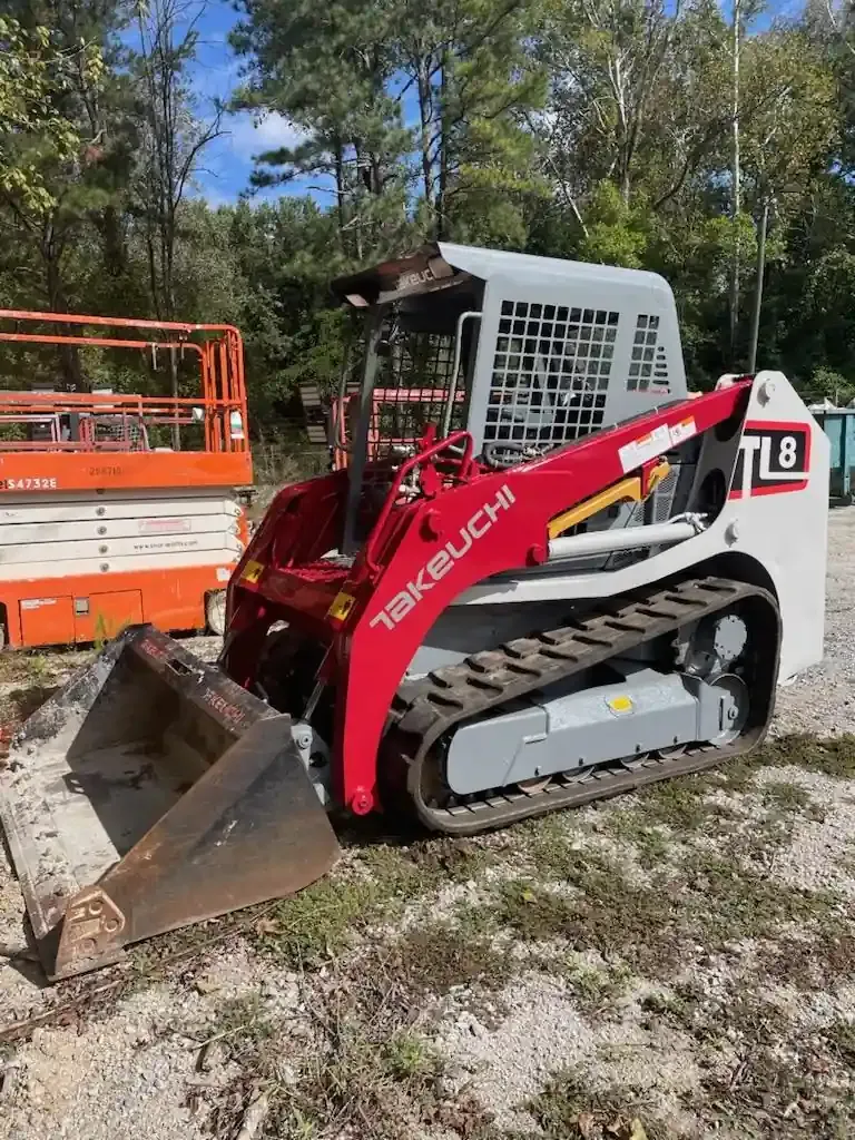 Red Takeuchi track loader with front bucket parked on dirt beside an orange trailer and trees