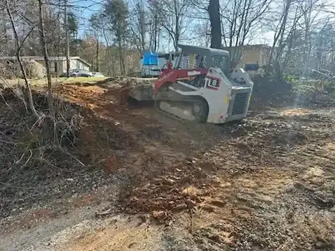 Compact skid steer clearing brush in a wooded lot near a house, with a blue dumpster in the background.