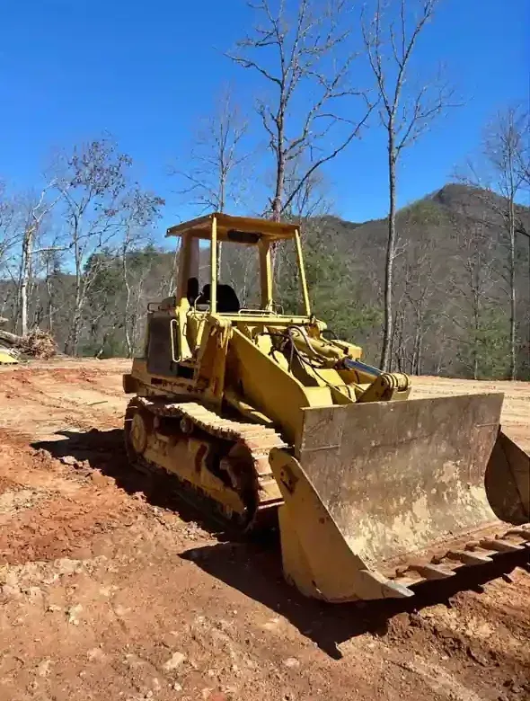 Yellow bulldozer on a dirt road in a wooded, mountainous area under a clear blue sky
