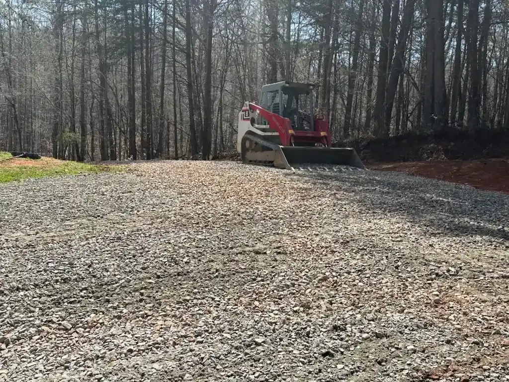 Red excavator on a gravel lot beside a wooded area in sunlight