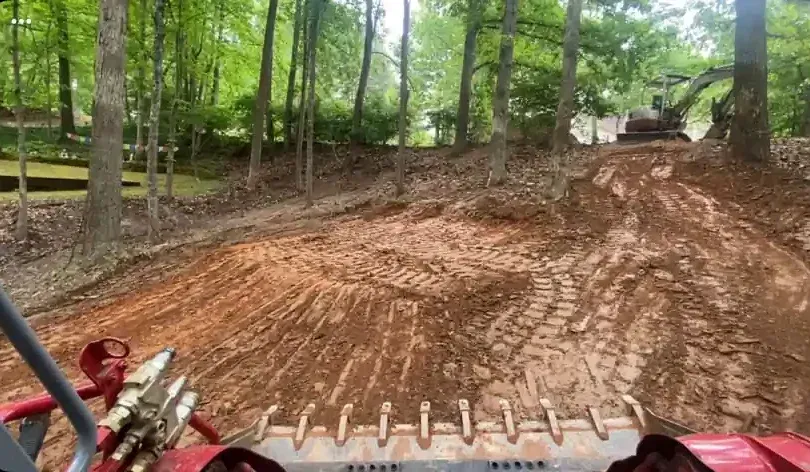 Dirt bike rider approaching a steep muddy hill in a wooded trail, with another bike visible at left