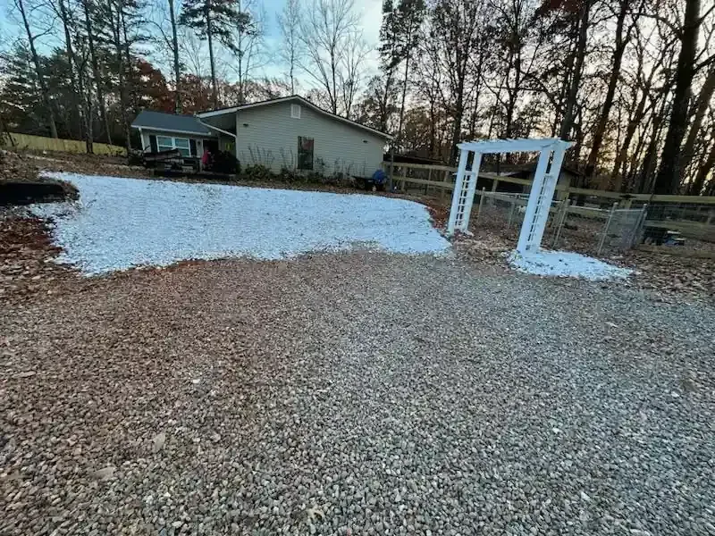 Gravel driveway with patches of snow and a white archway in front of a small house and trees