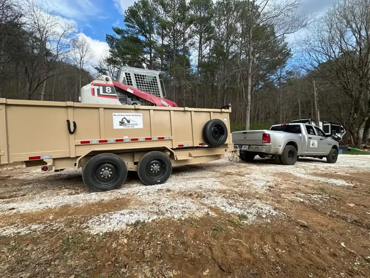 Towed tan trailer and silver pickup on a muddy lot with trees and a red crane in the background